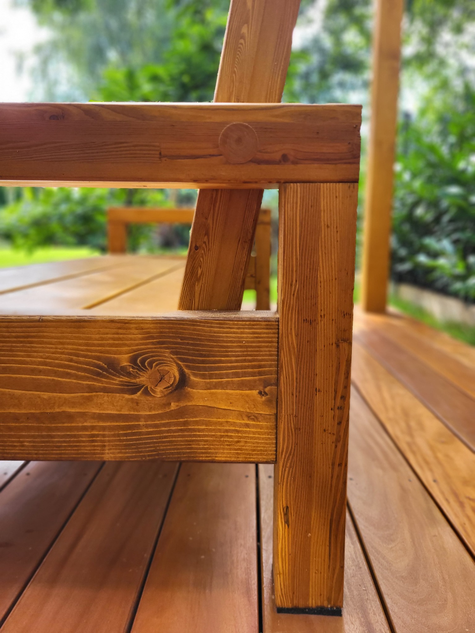 Fragment drewnianego tarasu z elementami mebli ogrodowych w ciepłym odcieniu brązu, z zielonym tłem roślinności. Widoczna struktura drewna i detale łączeń.