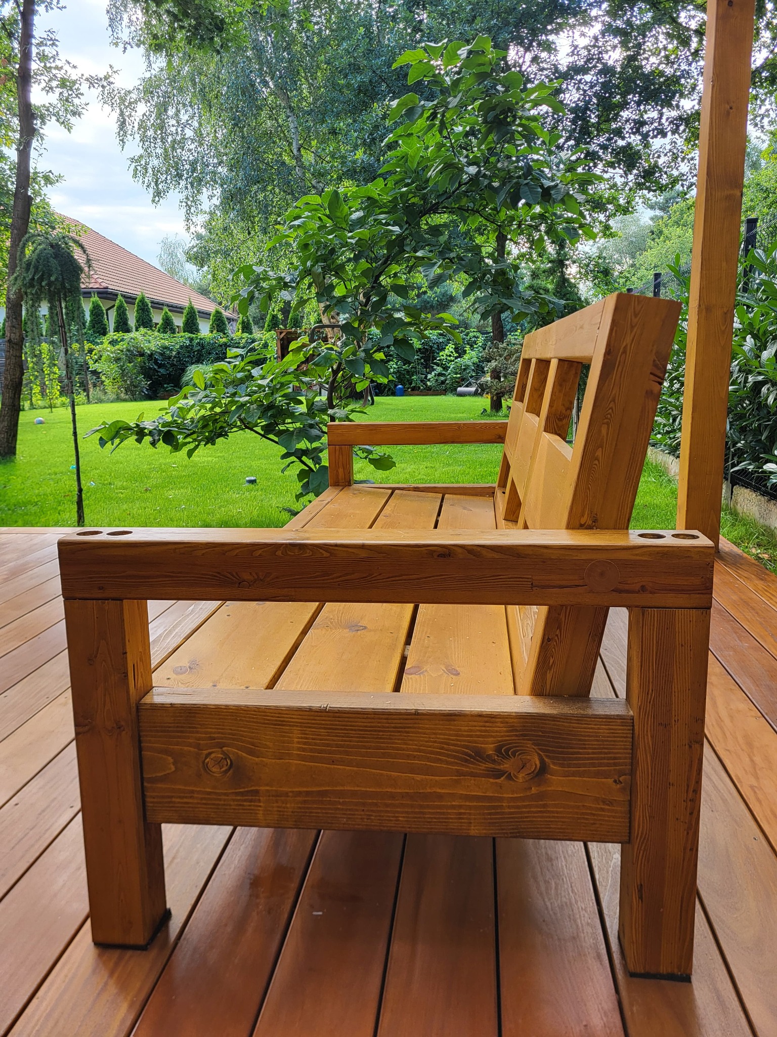 Drewniana ławka z uchwytami na napoje na tarasie z desek, w tle zielony trawnik i drzewa. Widok z boku, podkreślający solidną konstrukcję i naturalne wykończenie.