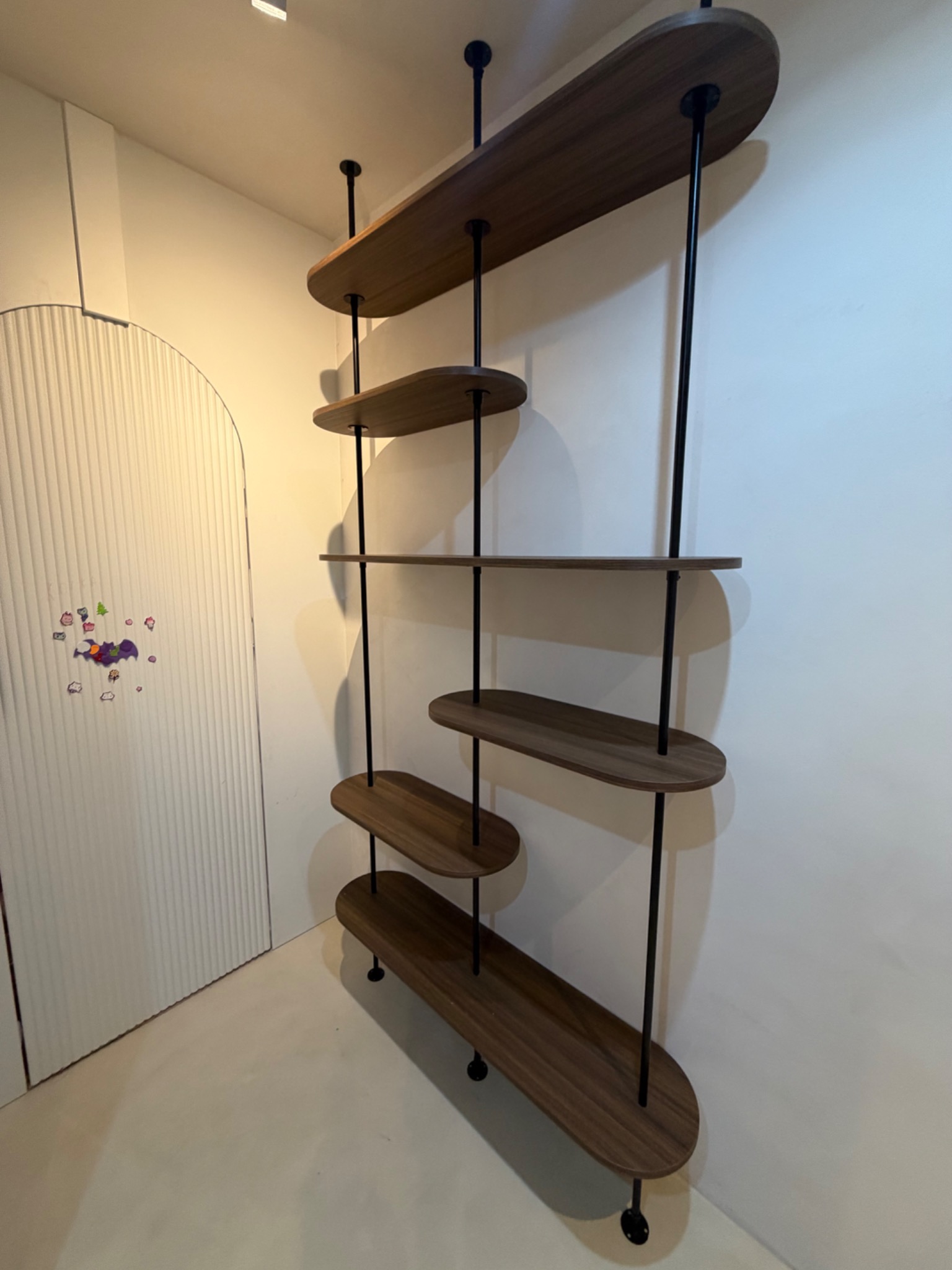 Nowoczesny regał z drewnianymi półkami o zaokrąglonym kształcie i czarnymi metalowymi wspornikami, stojący przy białej ścianie i drzwiach z dekoracyjnymi żłobieniami.