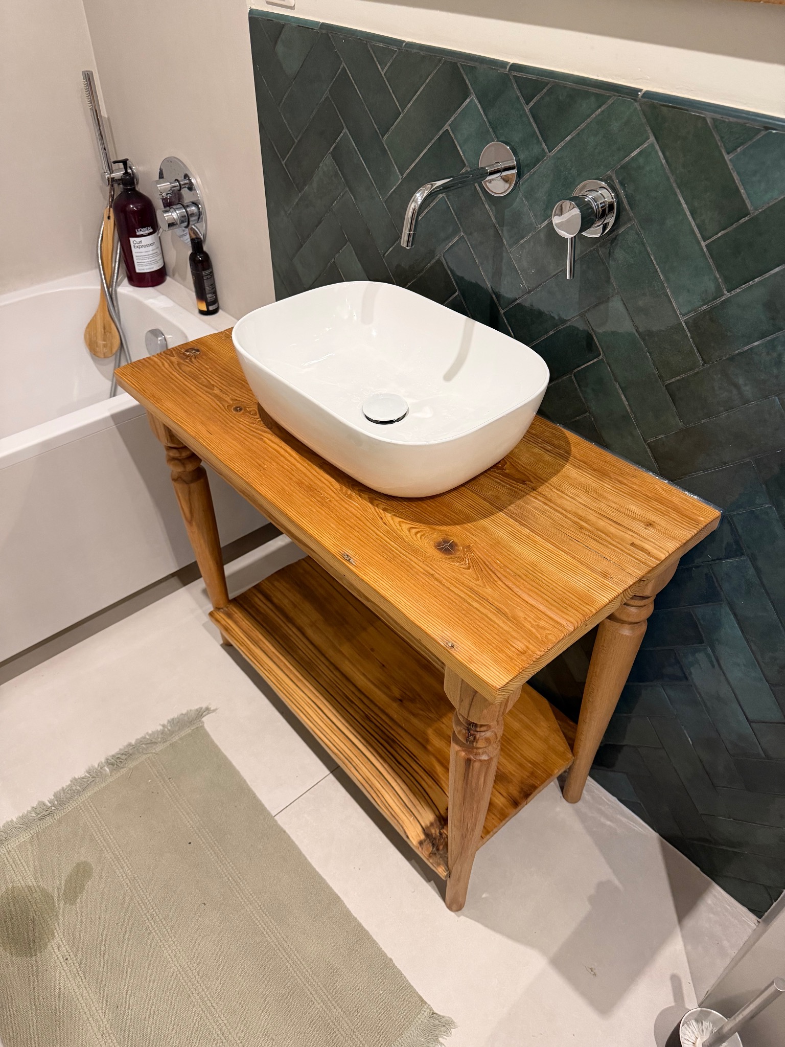 Biała umywalka nablatowa na drewnianym blacie w łazience z zielonymi płytkami ułożonymi w jodełkę. Widoczna bateria ścienna i fragment wanny.