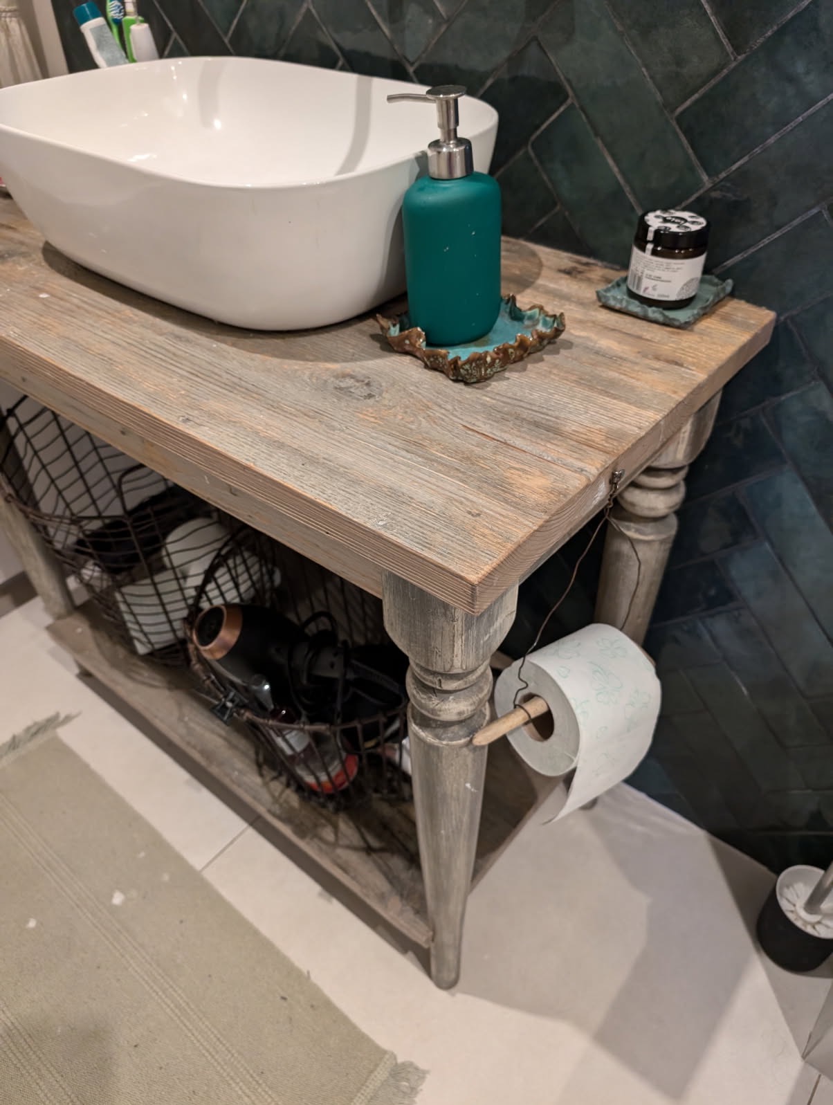 Rustykalna szafka łazienkowa z umywalką nablatową, dozownikiem mydła i koszem z akcesoriami. Uwagę przykuwa nietypowy uchwyt na papier toaletowy z drewnianym kołkiem.