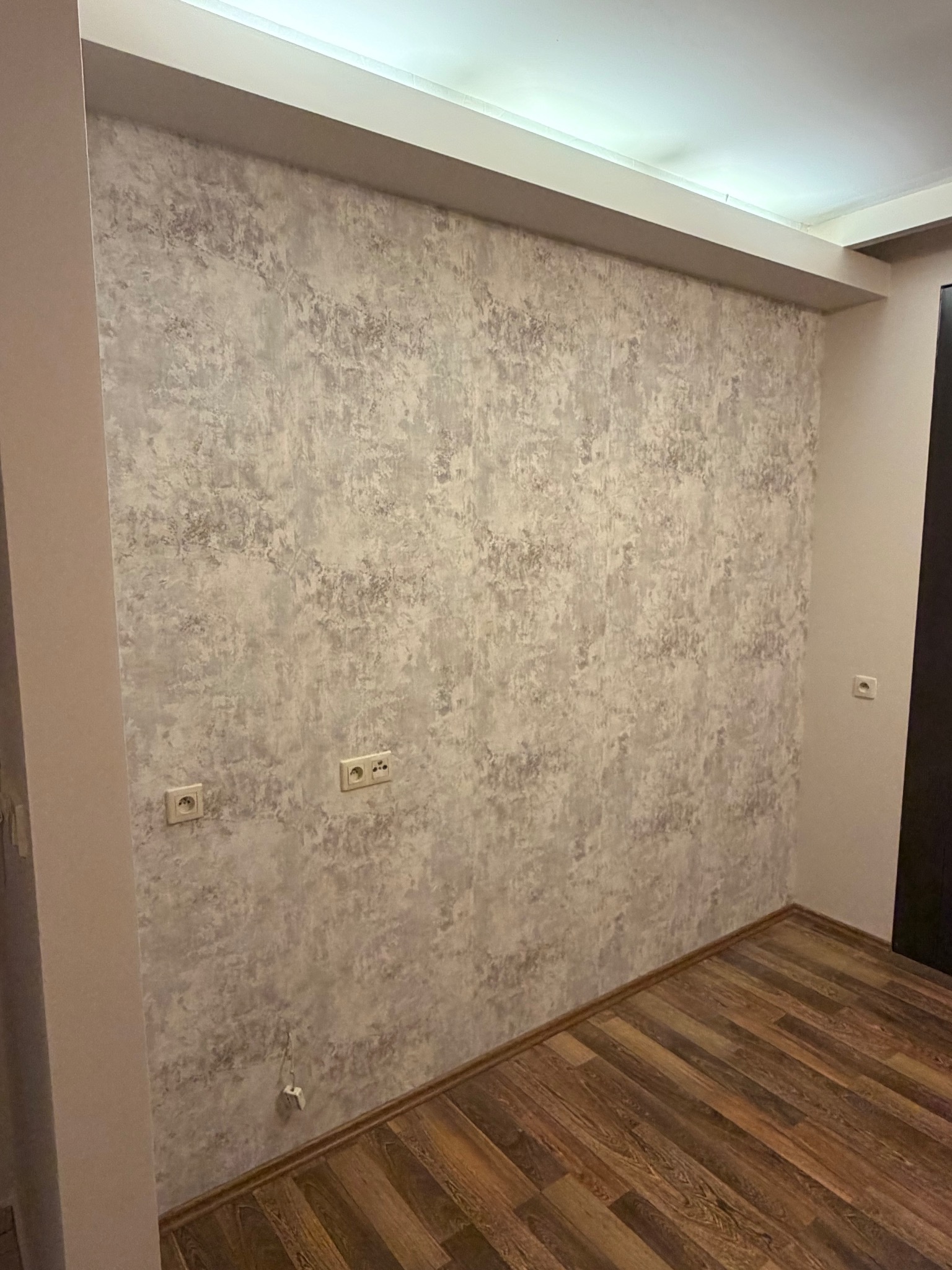 Ściana z dekoracyjną szarą tapetą o nieregularnym wzorze, widoczne gniazdka elektryczne i fragment drewnianej podłogi. Ujęcie z dołu, podkreślające fakturę ściany.