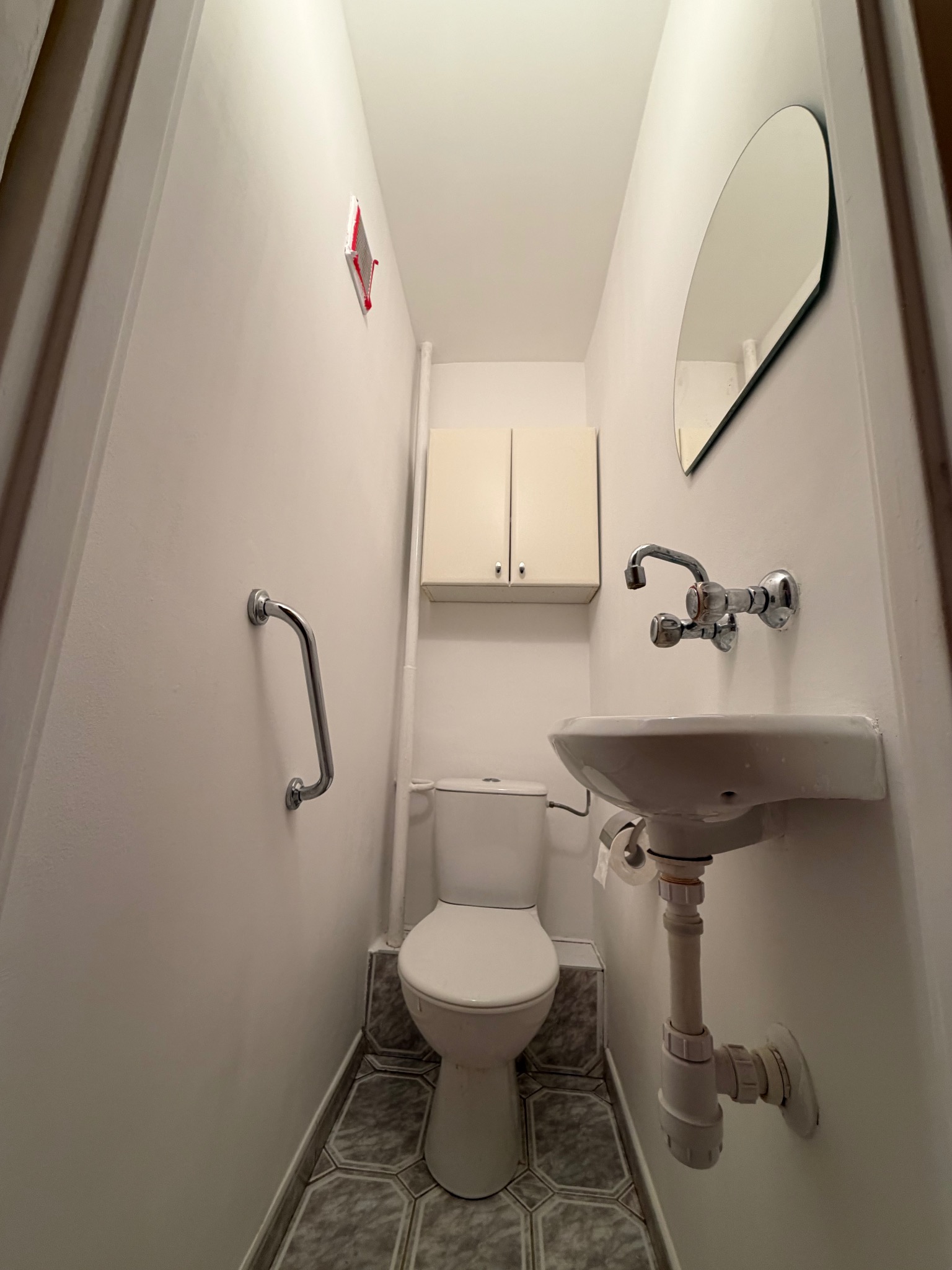 Wąska toaleta po remoncie: białe ściany, umywalka z lustrem, toaleta i szara podłoga z płytek. Dodatkowo zamontowany uchwyt dla niepełnosprawnych.