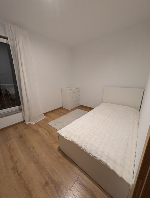 Jasne wnętrze sypialni po remoncie: drewniana podłoga, białe ściany, minimalistyczne meble i dywan. Widok na okno z białymi zasłonami.