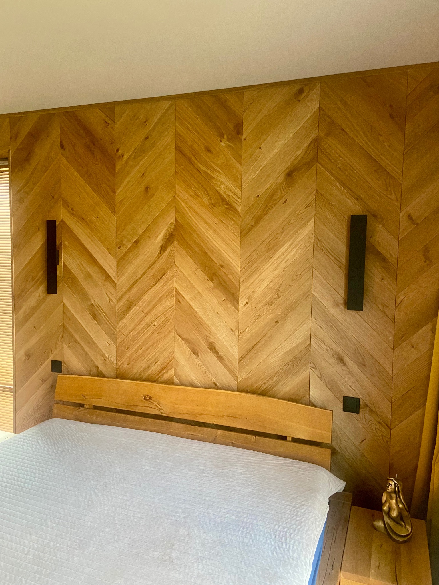 Drewniana ściana w jodełkę za łóżkiem, z minimalistycznymi lampami ściennymi. Widoczny fragment łóżka i stolika nocnego z dekoracyjną figurką.