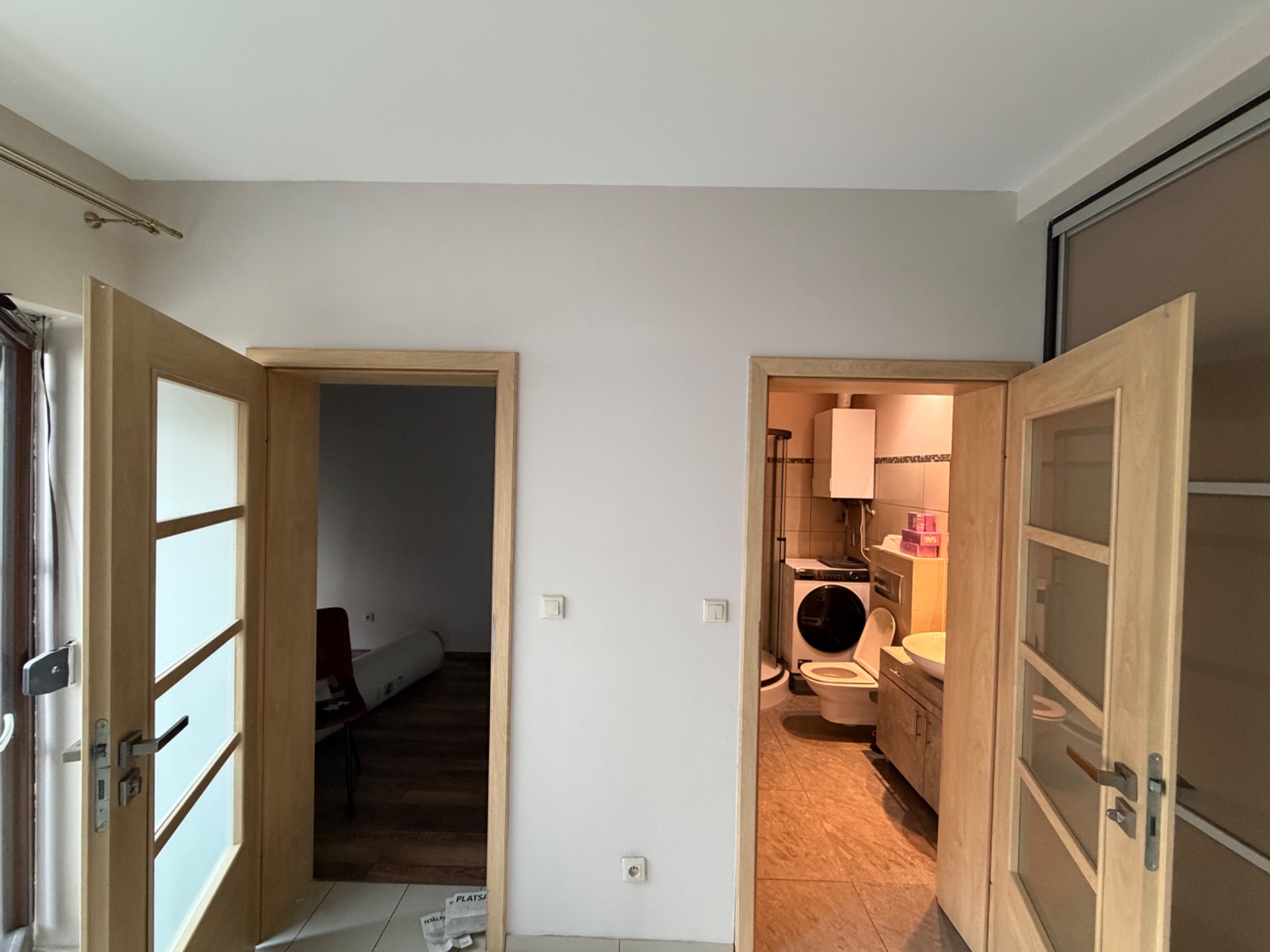 Dwa otwarte, drewniane drzwi z przeszkleniami we Wrocławiu, prowadzące do pokoju i łazienki. Świeżo pomalowane ściany, widoczne gniazdka elektryczne. Jasne wnętrze.