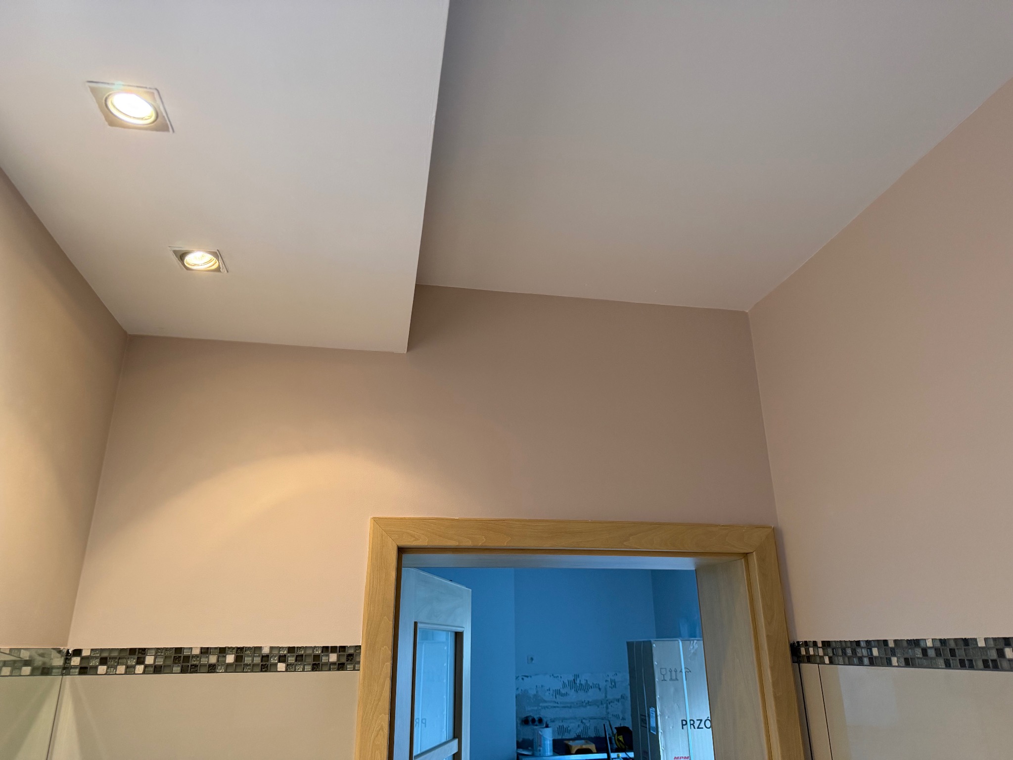 Pudrowo-różowa ściana po malowaniu z drewnianą ościeżnicą i mozaiką. Na suficie dwa kwadratowe reflektory. Widok na sąsiednie pomieszczenie przez otwarte drzwi.