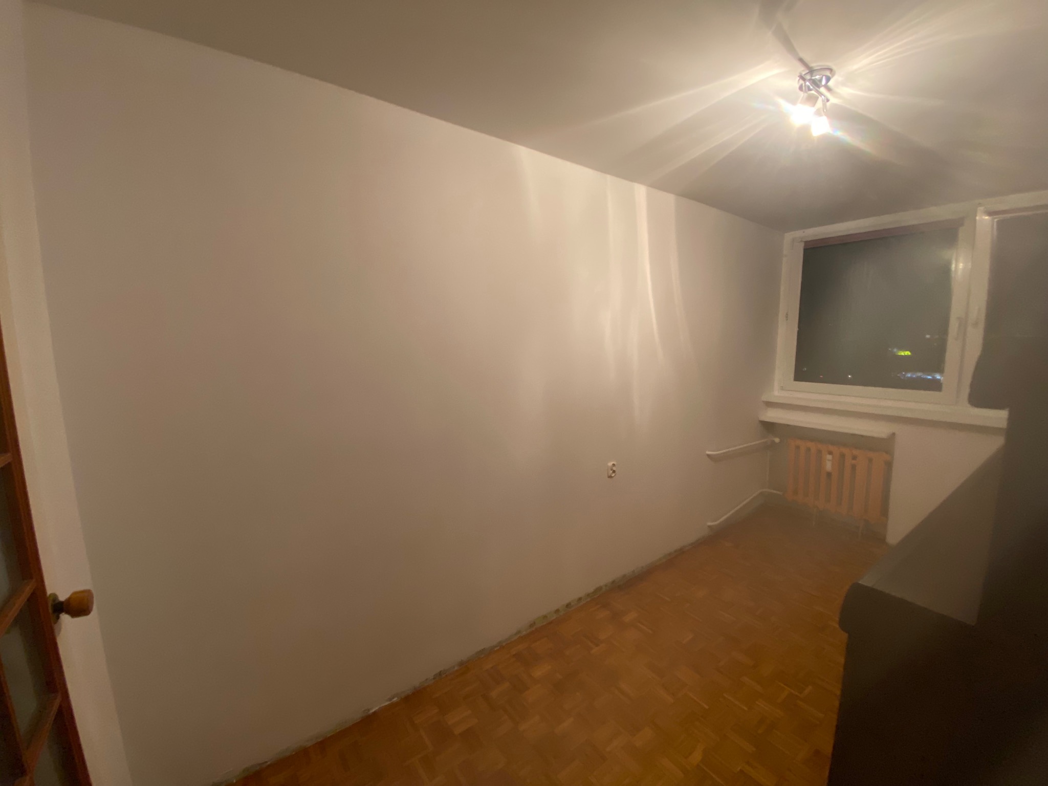 Pomalowane na biało wnętrze pokoju z parkietem, grzejnikiem pod oknem i widocznym fragmentem drzwi po lewej stronie. Świeże, jasne ściany po malowaniu.