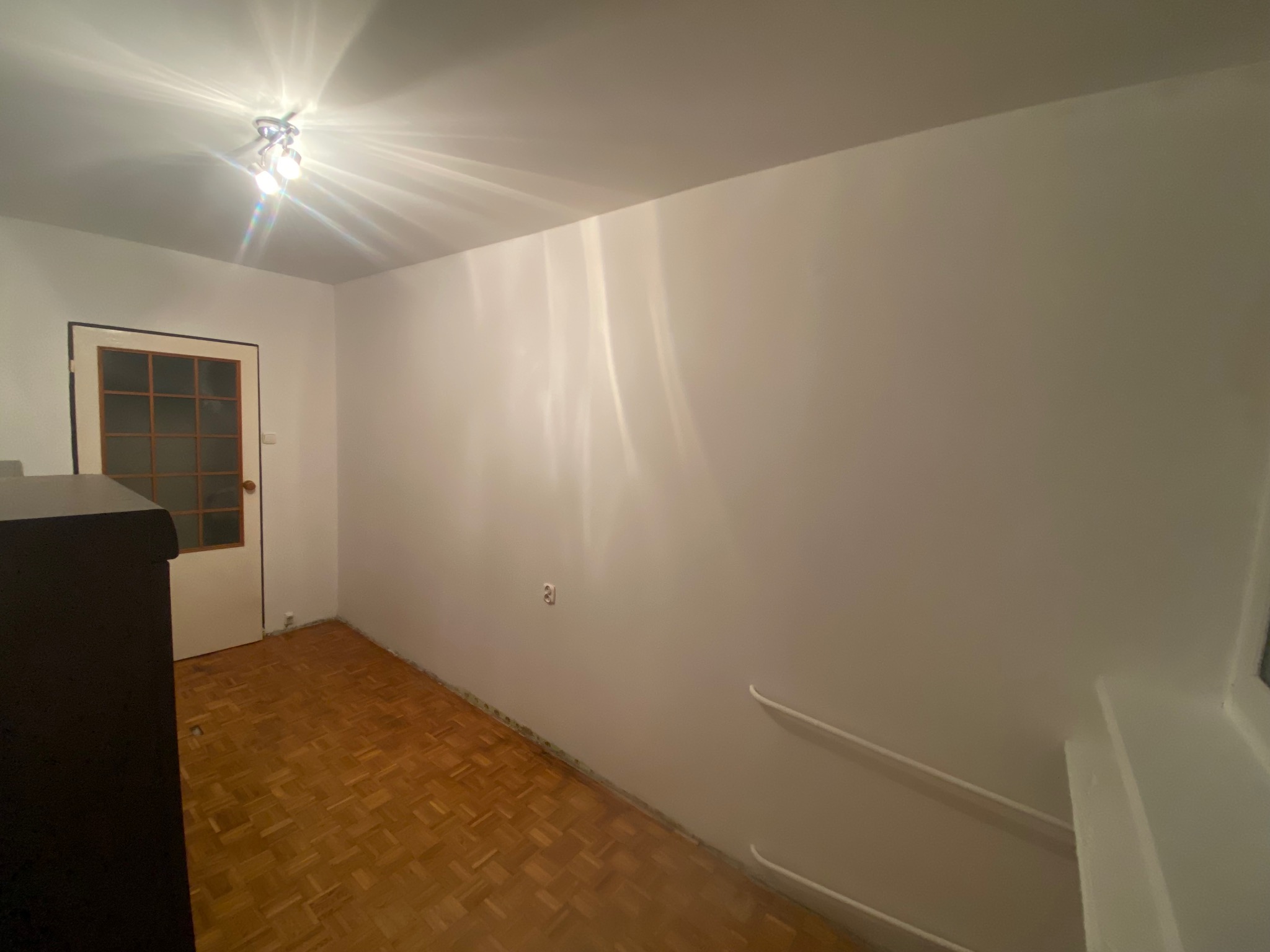 Jasne wnętrze z parkietem w jodełkę, świeżo pomalowane ściany i białe listwy przypodłogowe. Widoczna lampa sufitowa i drzwi z szybami.