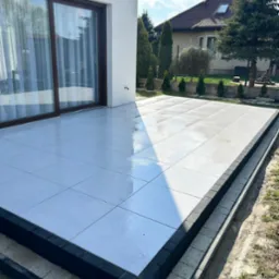 Nowoczesny taras z jasnych płyt, obrzeża z ciemnej kostki. Widok na dom i ogród w słoneczny dzień.