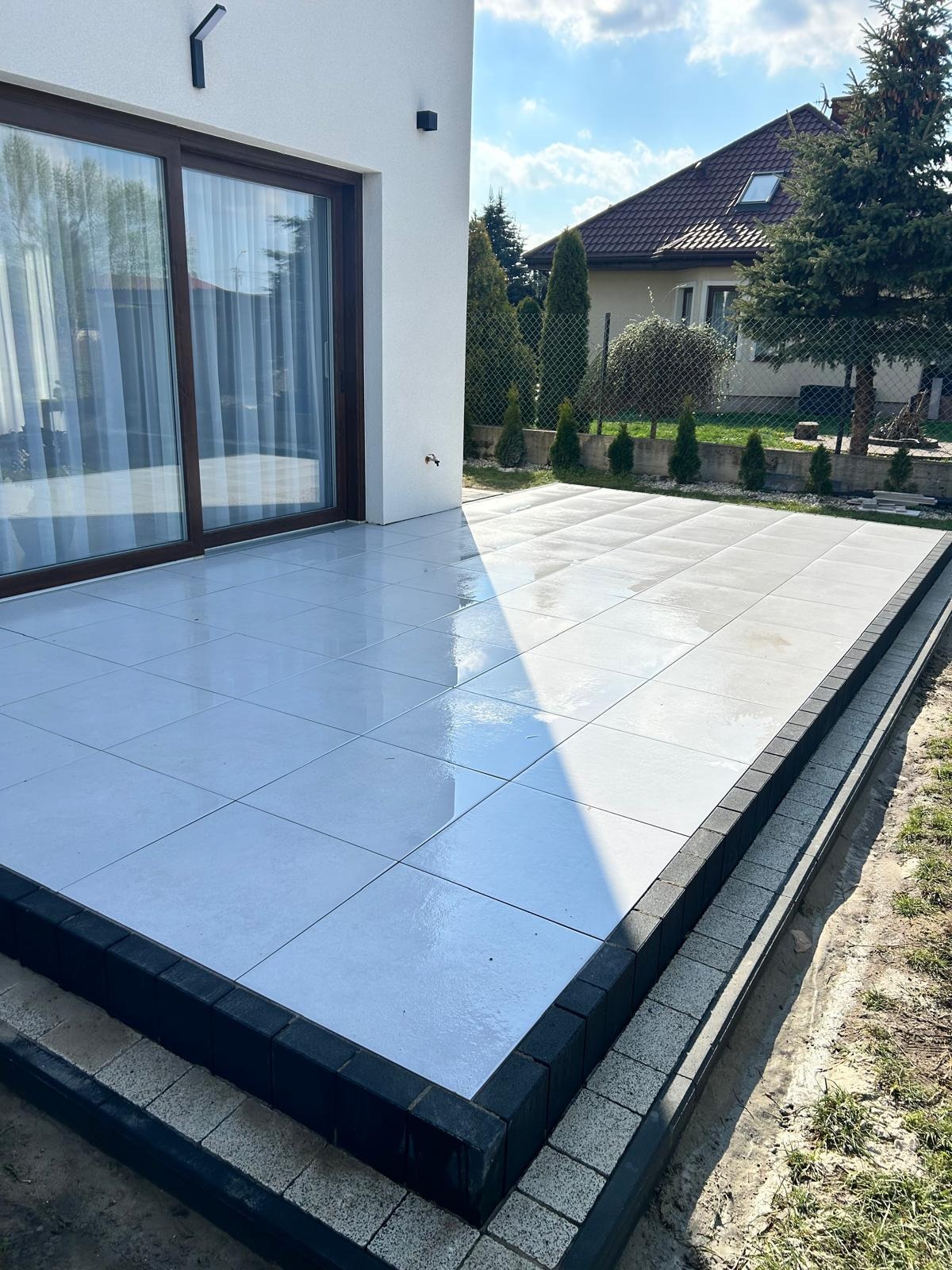 Nowoczesny taras z jasnych płyt, obrzeża z ciemnej kostki. Widok na dom i ogród w słoneczny dzień.