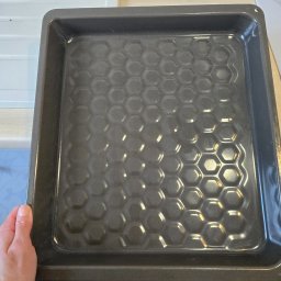 Angelika Kowal - Brudna blacha do pieczenia z tłustymi śladami w kuchni, trzymana przez osobę. Widoczne wnętrze piekarnika i fragment zlewu. Blacha z wzorem w kształcie plastra miodu.