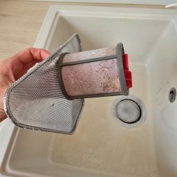 Angelika Kowal - Zanieczyszczony filtr okapu kuchennego trzymany nad zlewem, gotowy do umycia. Widoczne resztki tłuszczu i zabrudzeń. Metalowa siatka filtra o drobnych oczkach.