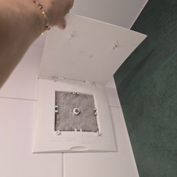 Angelika Kowal - Otwarty, biały panel dostępowy z szarym filtrem wewnątrz, prawdopodobnie wentylacyjny, na tle białych i zielonych płytek. Widoczna ręka z bransoletką.