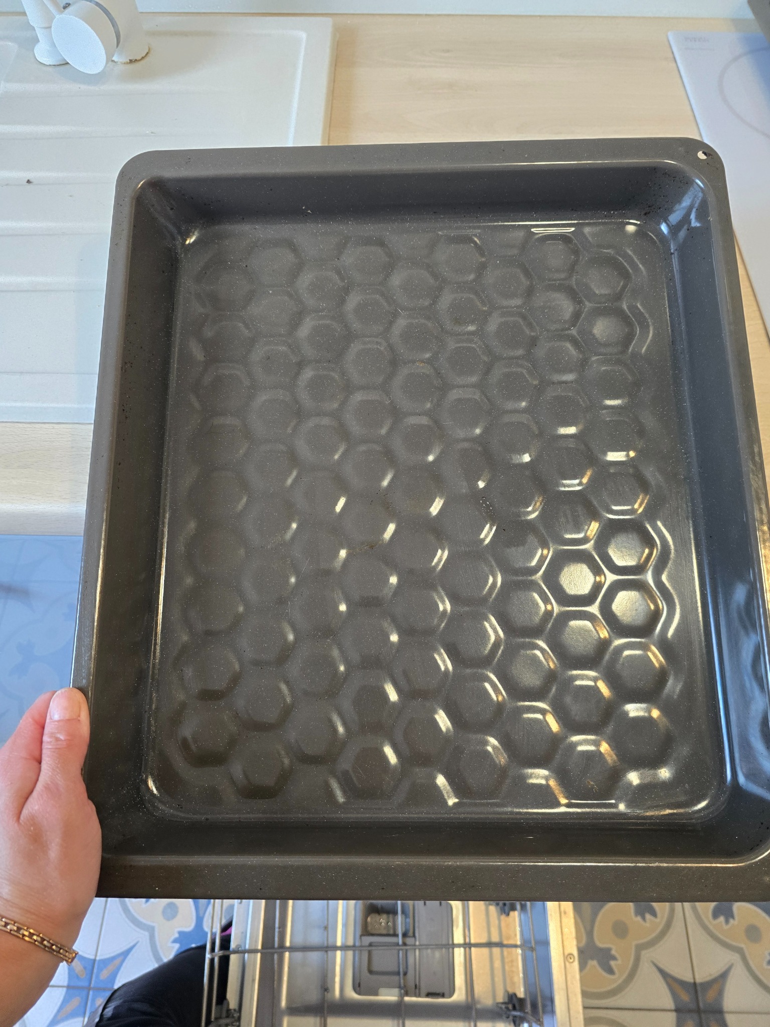 Brudna blacha do pieczenia z tłustymi śladami w kuchni, trzymana przez osobę. Widoczne wnętrze piekarnika i fragment zlewu. Blacha z wzorem w kształcie plastra miodu.