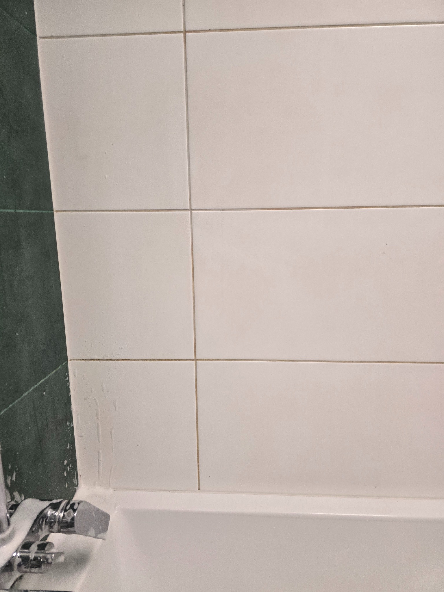 Białe płytki w łazience z zielonym akcentem, widoczny kran z pianą, sugerujący czyszczenie. Ujęcie z bliska, skupione na fugach i powierzchniach.