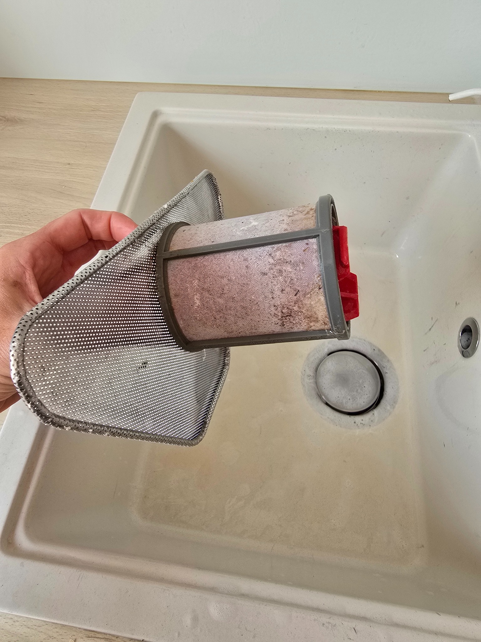 Zanieczyszczony filtr okapu kuchennego trzymany nad zlewem, gotowy do umycia. Widoczne resztki tłuszczu i zabrudzeń. Metalowa siatka filtra o drobnych oczkach.