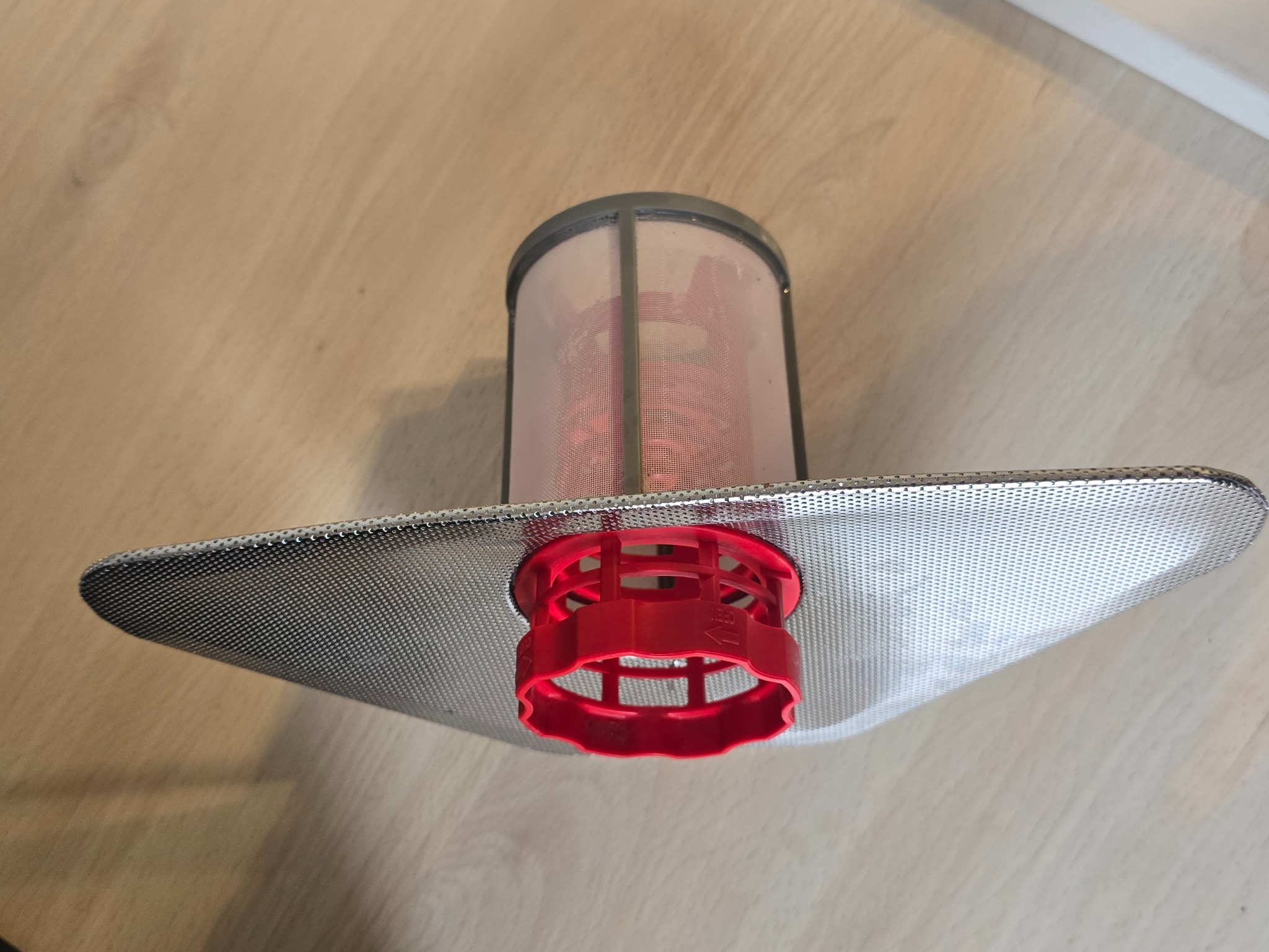 Filtr do zmywarki z metalową siatką i czerwonym elementem z tworzywa sztucznego, leżący na drewnianej powierzchni. Widoczne ślady użytkowania.