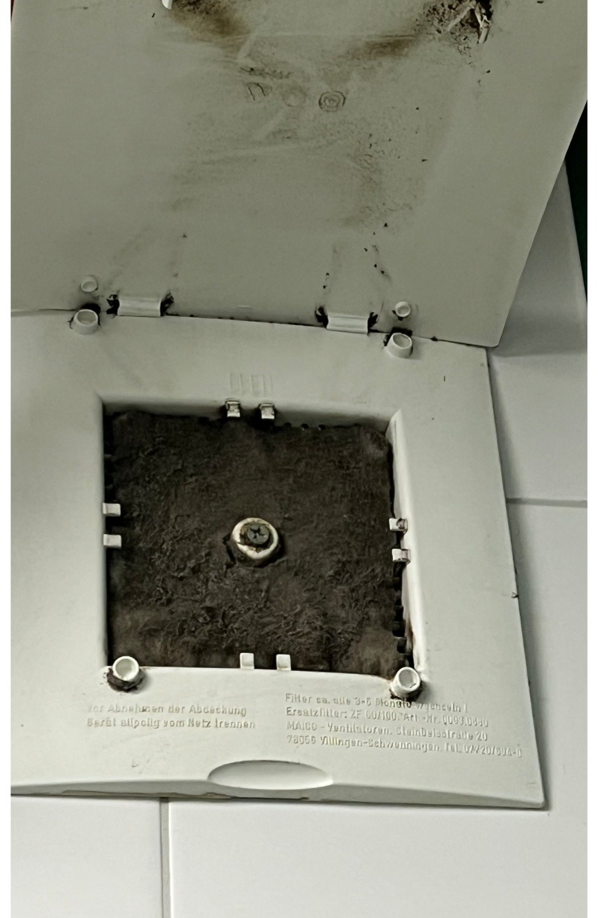 Zanieczyszczony filtr wentylacyjny w białej obudowie, widoczne nagromadzenie kurzu i brudu. Filtr wymaga wymiany lub czyszczenia.