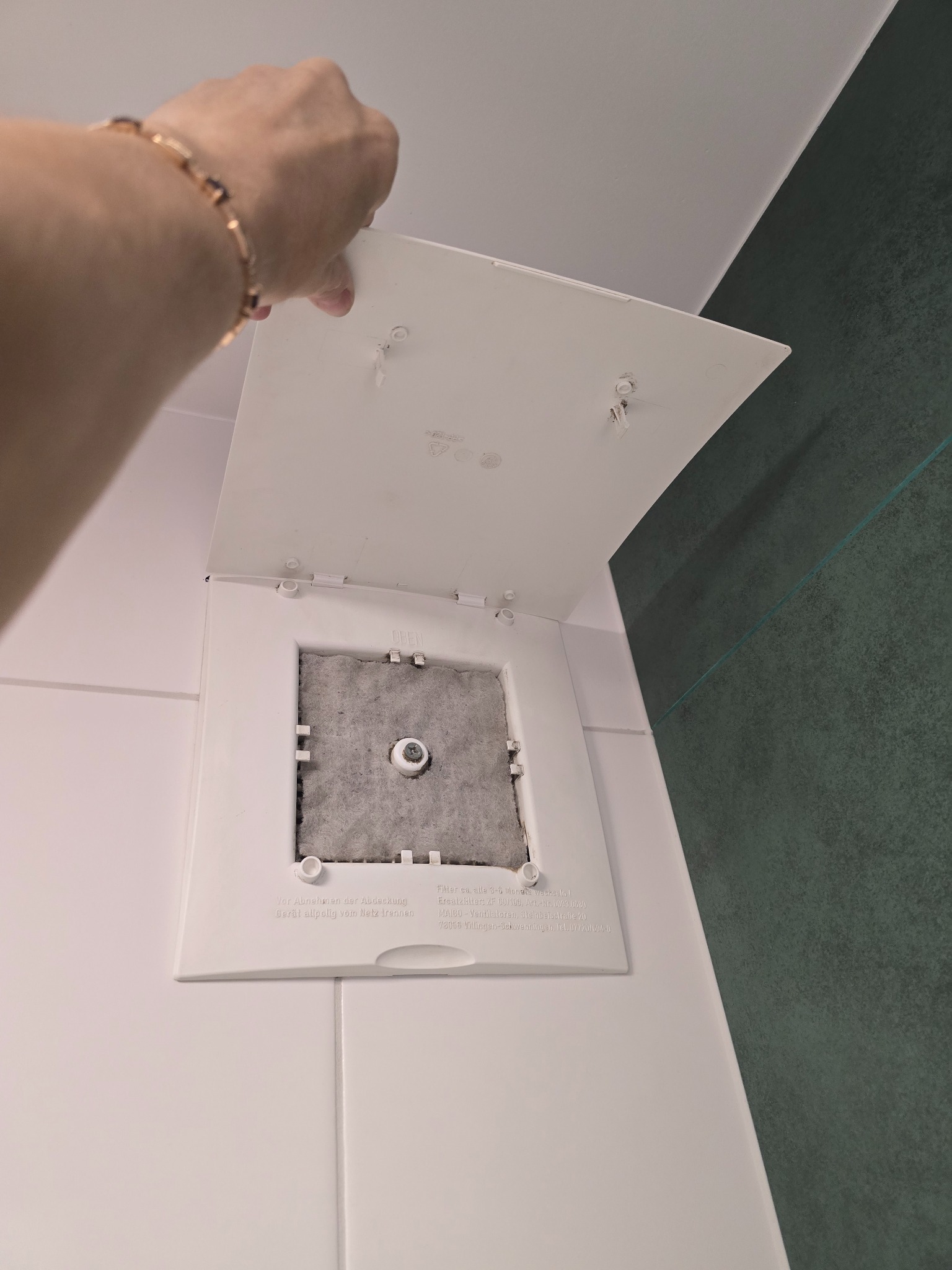 Otwarty, biały panel dostępowy z szarym filtrem wewnątrz, prawdopodobnie wentylacyjny, na tle białych i zielonych płytek. Widoczna ręka z bransoletką.