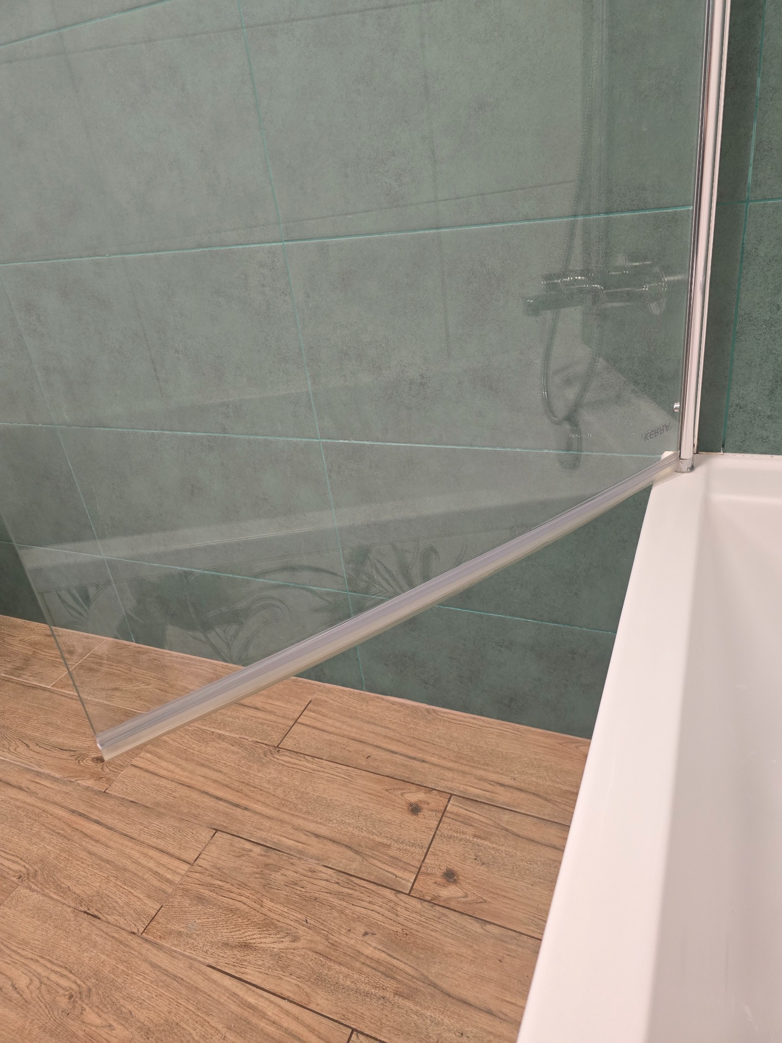 Szklana kabina prysznicowa z chromowanymi elementami, zielone płytki na ścianie i drewnopodobna podłoga. Widok na fragment wanny i baterię.