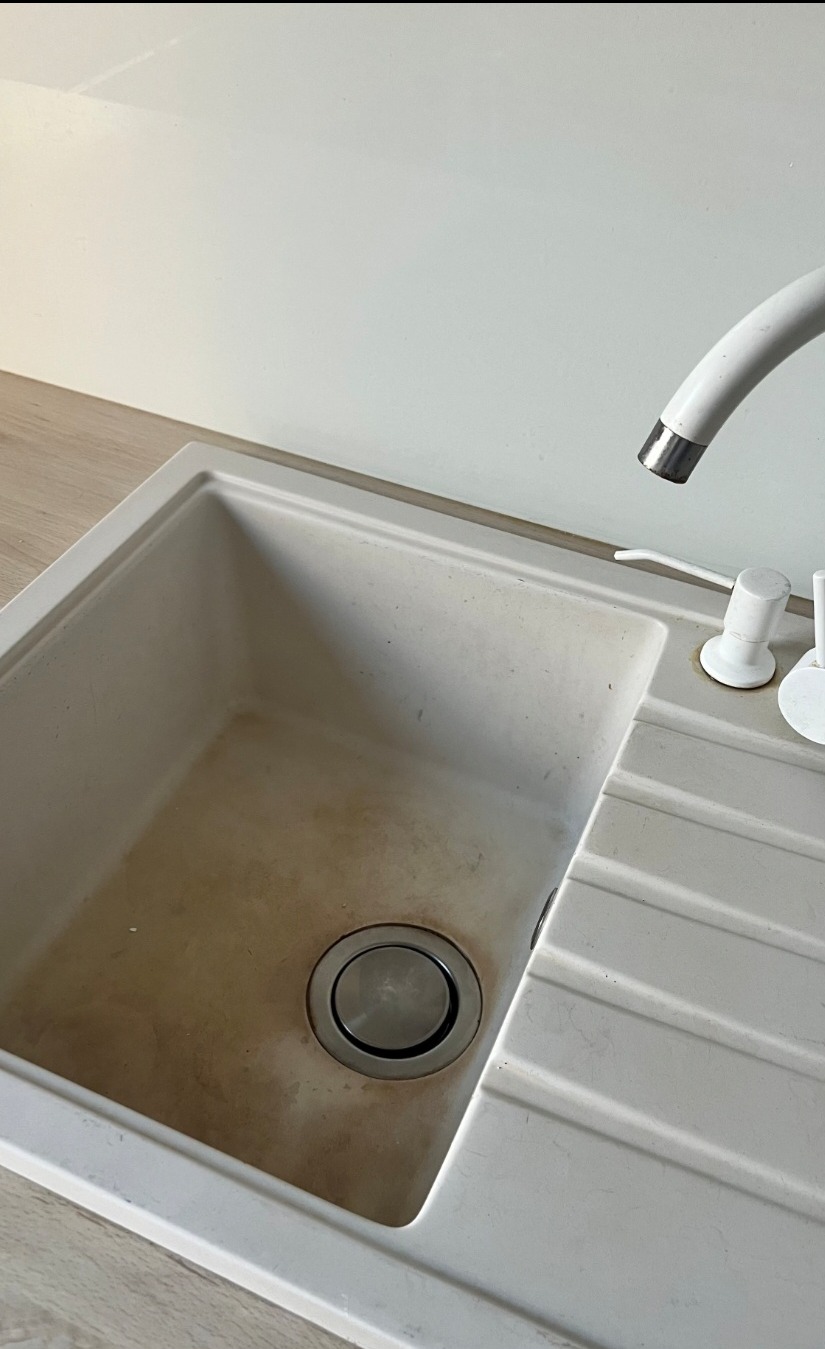 Zabrudzony, biały zlewozmywak granitowy z ociekaczem i kranem. Widoczne ślady użytkowania i osady. Ujęcie z góry, z bliska.