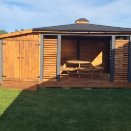 Pasjion wood MAŁGORZATA ADRYCH - Drewniana altana z zamykaną częścią i stołem piknikowym, otoczona zielonym trawnikiem i kamieniami. Konstrukcja pomalowana na brązowo i szaro, z czarnym dachem.