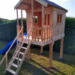 Drewniany domek zabaw dla dzieci z niebieską zjeżdżalnią i schodami, ustawiony na trawniku przed zielonym żywopłotem. Naturalne drewno, prosta konstrukcja.