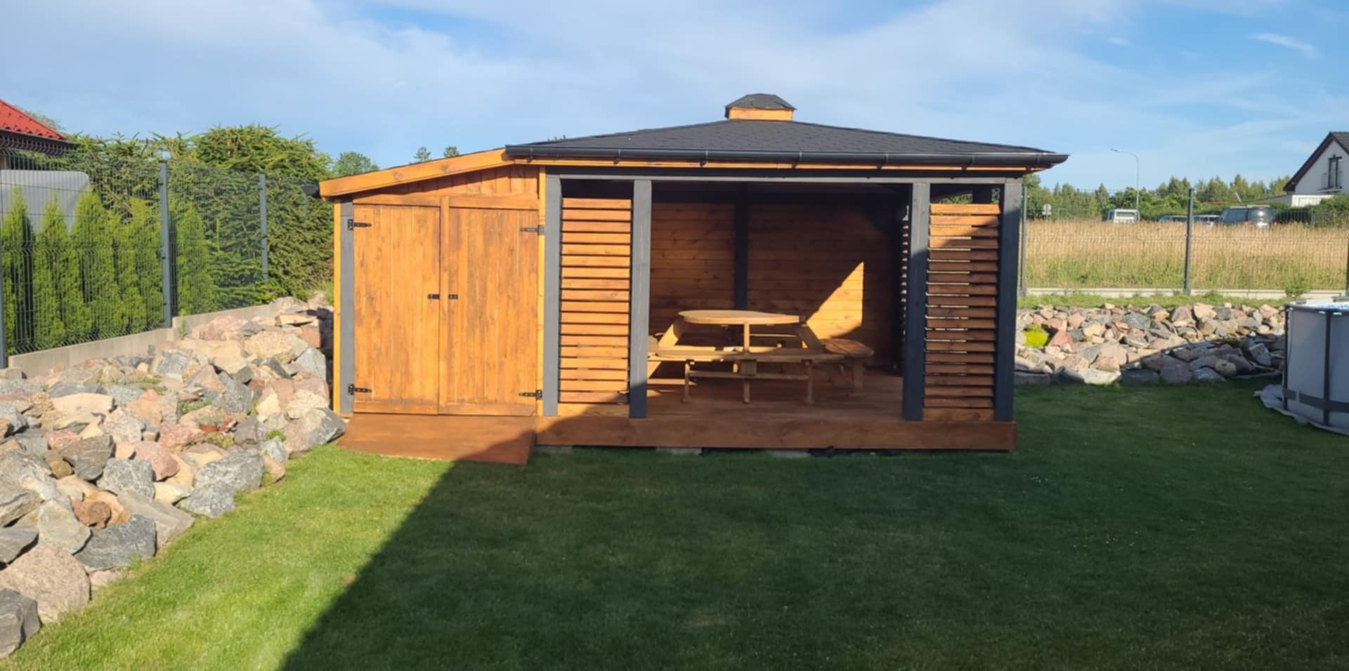 Drewniana altana z zamykaną częścią i stołem piknikowym, otoczona zielonym trawnikiem i kamieniami. Konstrukcja pomalowana na brązowo i szaro, z czarnym dachem.