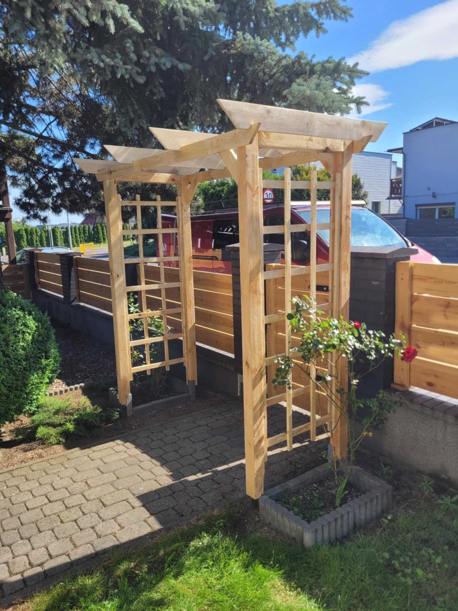 Surowa, drewniana pergola z kratownicą, z pnącą różą, na tle ogrodzenia z drewnianymi panelami. Słoneczny dzień, zielony trawnik i krzewy.