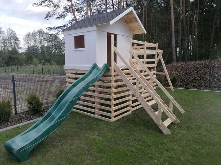 Drewniany domek zabaw dla dzieci z zieloną zjeżdżalnią i schodami na trawniku, otoczony siatką ogrodzeniową i zielenią. Naturalne otoczenie.