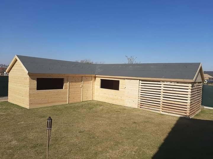 Kątowa, drewniana altana z szarą dachówką, oknami i ażurową ścianą, stojąca na trawniku w słoneczny dzień. W tle widoczne domy i zielone ogrodzenie.