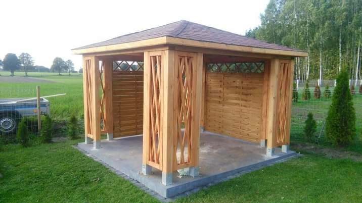 Drewniana altana z dekoracyjnymi kolumnami i ażurowymi ścianami w Niedźwiedziu, na tle zielonego trawnika i drzew. Widoczny fragment przyczepy.