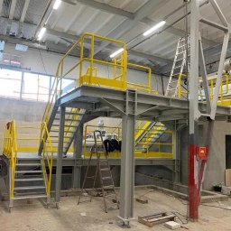 STEEL-TECH SERVICES SPÓŁKA Z OGRANICZONĄ ODPOWIEDZIALNOŚCIĄ - Wnętrze hali z metalową konstrukcją schodów i podestów w szaro-żółtej kolorystyce. Widoczne drabiny, okno i elementy instalacji pod sufitem.