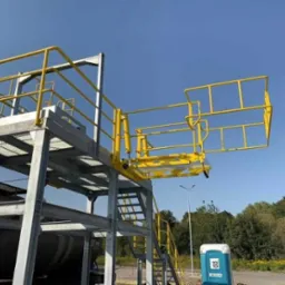 Stalowa konstrukcja z żółtymi poręczami, schodami i wysuwaną platformą roboczą na tle błękitnego nieba. Widoczny fragment zbiornika i toaleta Toi Toi.