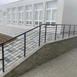 STEEL-TECH SERVICES SPÓŁKA Z OGRANICZONĄ ODPOWIEDZIALNOŚCIĄ - Zewnętrzna, czarna, metalowa balustrada schodowa z poziomymi prętami, zamontowana przy budynku w pochmurny dzień. Widoczne szare płytki chodnikowe i schody.