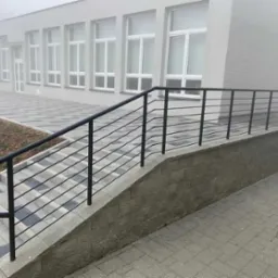 Zewnętrzna, czarna, metalowa balustrada schodowa z poziomymi prętami, zamontowana przy budynku w pochmurny dzień. Widoczne szare płytki chodnikowe i schody.