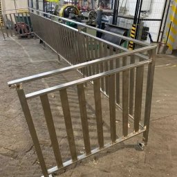 STEEL-TECH SERVICES SPÓŁKA Z OGRANICZONĄ ODPOWIEDZIALNOŚCIĄ - Nierdzewna balustrada schodowa z pionowymi szczeblami, w surowym wnętrzu hali produkcyjnej. Widoczne ślady obróbki metalu i elementy wyposażenia warsztatu.