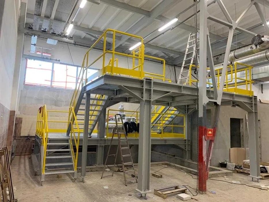 Wnętrze hali z metalową konstrukcją schodów i podestów w szaro-żółtej kolorystyce. Widoczne drabiny, okno i elementy instalacji pod sufitem.