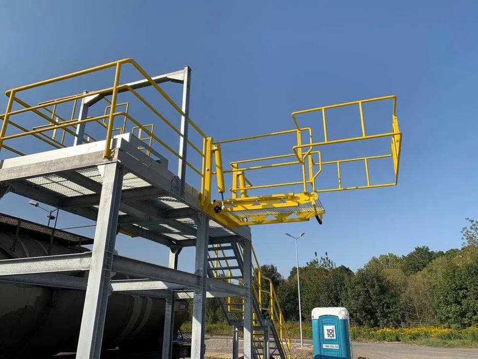 Stalowa konstrukcja z żółtymi poręczami, schodami i wysuwaną platformą roboczą na tle błękitnego nieba. Widoczny fragment zbiornika i toaleta Toi Toi.
