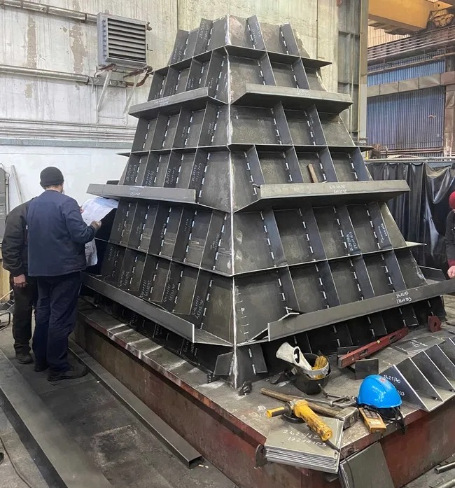 Konstrukcja stalowa w kształcie piramidy, z widocznymi spawami i osobami analizującymi plan. Na stole roboczym narzędzia, kask i poziomica.