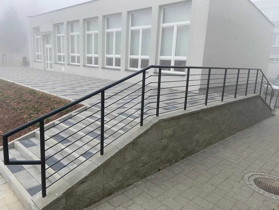 Zewnętrzna, czarna, metalowa balustrada schodowa z poziomymi prętami, zamontowana przy budynku w pochmurny dzień. Widoczne szare płytki chodnikowe i schody.