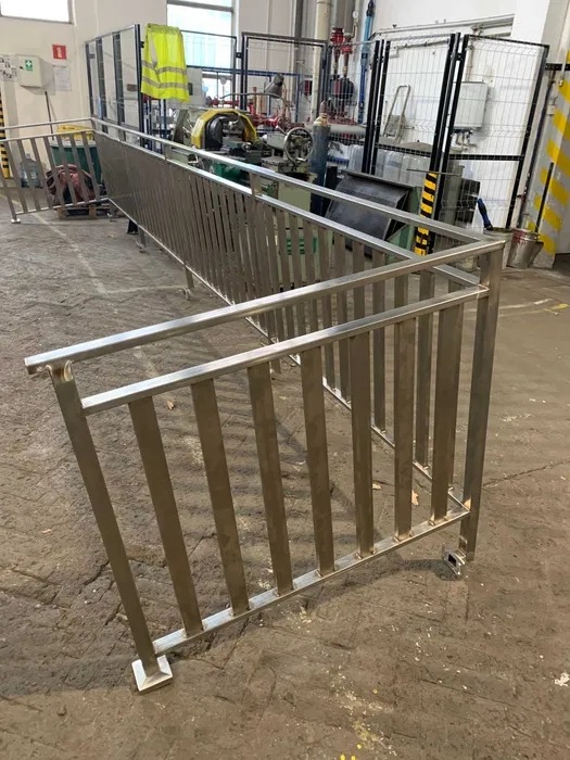 Nierdzewna balustrada schodowa z pionowymi szczeblami, w surowym wnętrzu hali produkcyjnej. Widoczne ślady obróbki metalu i elementy wyposażenia warsztatu.