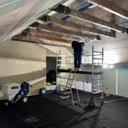 Prace przygotowawcze przed natryskiem gładzi w Żukowie. Widoczny pracownik na rusztowaniu, zabezpieczone elementy konstrukcyjne i agregat do gładzi.