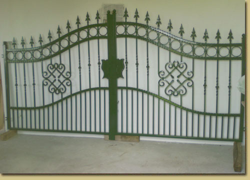 Zielona, metalowa brama wjazdowa z dekoracyjnymi elementami w kształcie serc i grotów, zamontowana tymczasowo na drewnianych klockach.