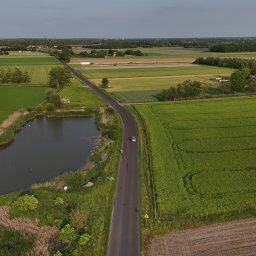 WOZelse - Bartosz Woźniak - Szeroki kadr z drona: wiejska droga przecina zielone pola uprawne, obok staw z trzciną, w oddali zabudowania. Samochód na drodze, znak ostrzegawczy.