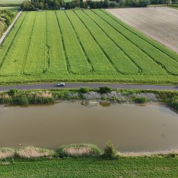 WOZelse - Bartosz Woźniak - Panoramiczny widok z drona na krajobraz rolniczy z polami, stawem i drogą, wzdłuż której jadą dwa samochody. Zielone pola i spokojna woda tworzą harmonijną scenerię.