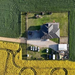 WOZelse - Bartosz Woźniak - Zdjęcie z drona: nowoczesny dom z ciemnym dachem otoczony zielonym i żółtym polem, liczne samochody na podjeździe, symetryczny układ kompozycyjny.