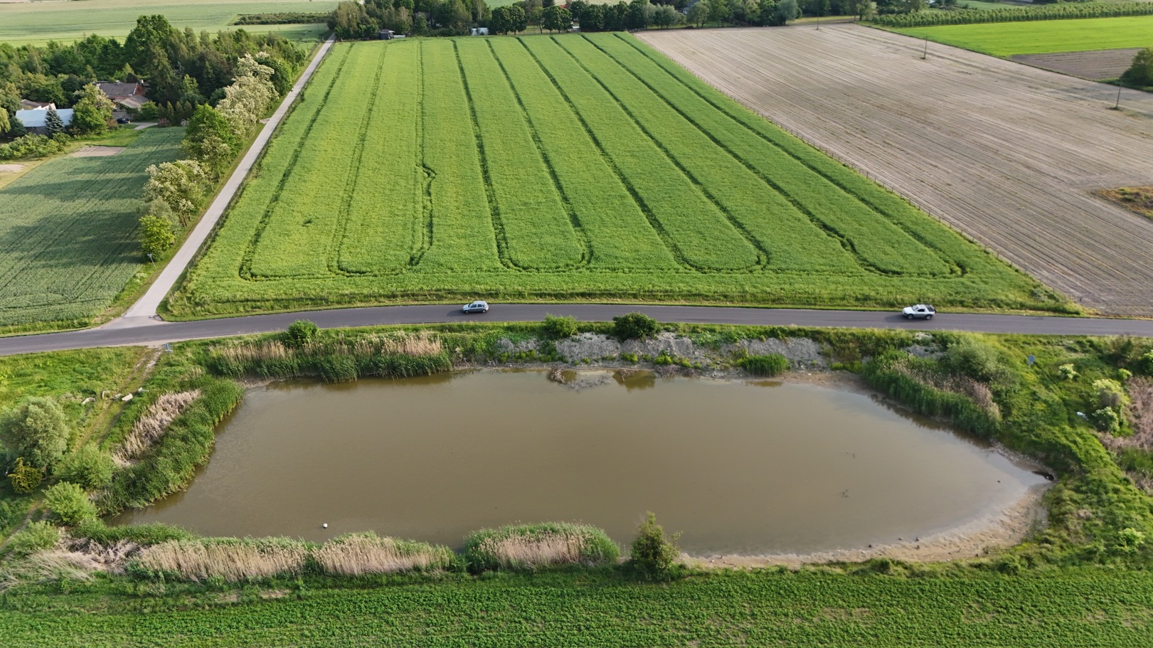 Panoramiczny widok z drona na krajobraz rolniczy z polami, stawem i drogą, wzdłuż której jadą dwa samochody. Zielone pola i spokojna woda tworzą harmonijną scenerię.
