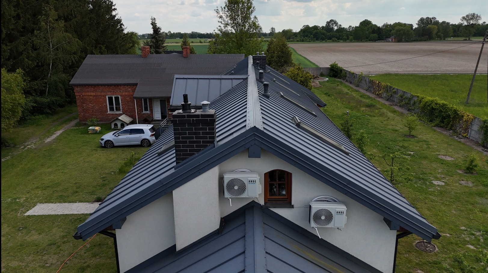 Metalowy dach z kominami i klimatyzatorami na tle zielonego ogrodu i pola. Widok z góry ukazuje szczegóły konstrukcji i otoczenie domu.
